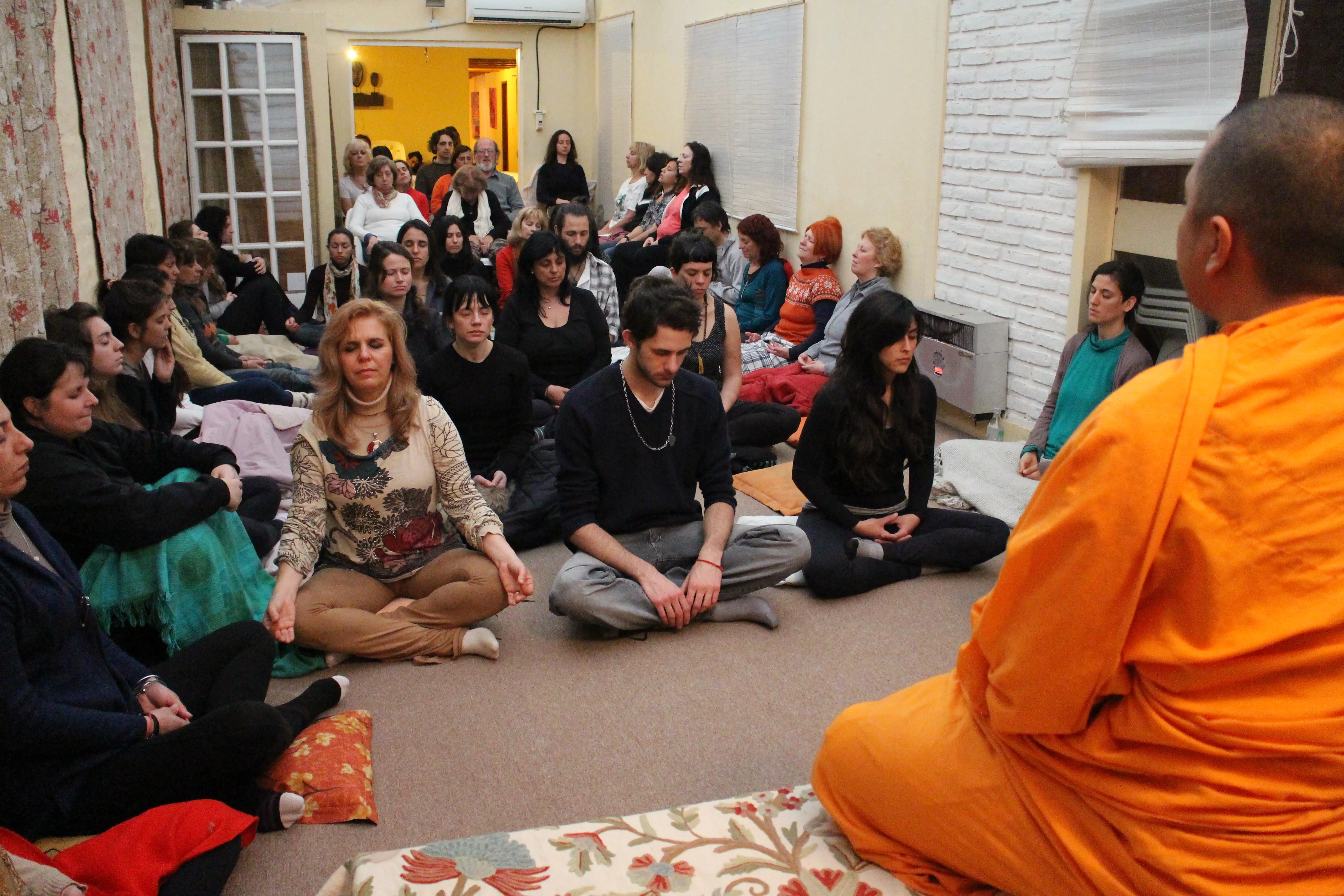 A crowded room sitting on the floor at Centro Shakti, facing a monk
